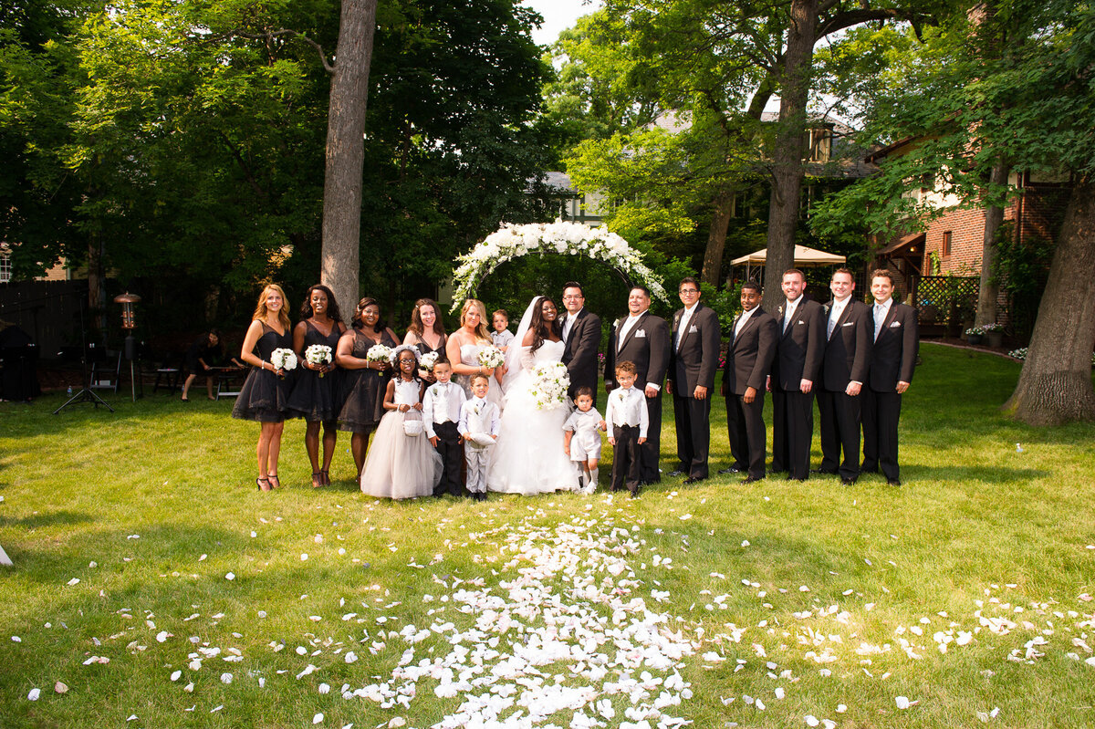 Newly wedded couple with guests beside in an outdoor ceremony setting with floral arch and flower petals on the ground