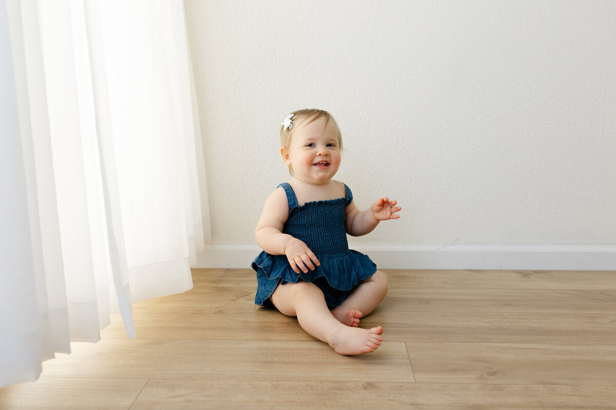 Adorable one-year-old girl sitting on the floor, wearing a charming denim dress, with a big smile on her face. The soft, natural light from the window highlights the pure joy of this sweet moment, captured by Yucaipa cake smash photographer Kaitlyn Dawn Photography.