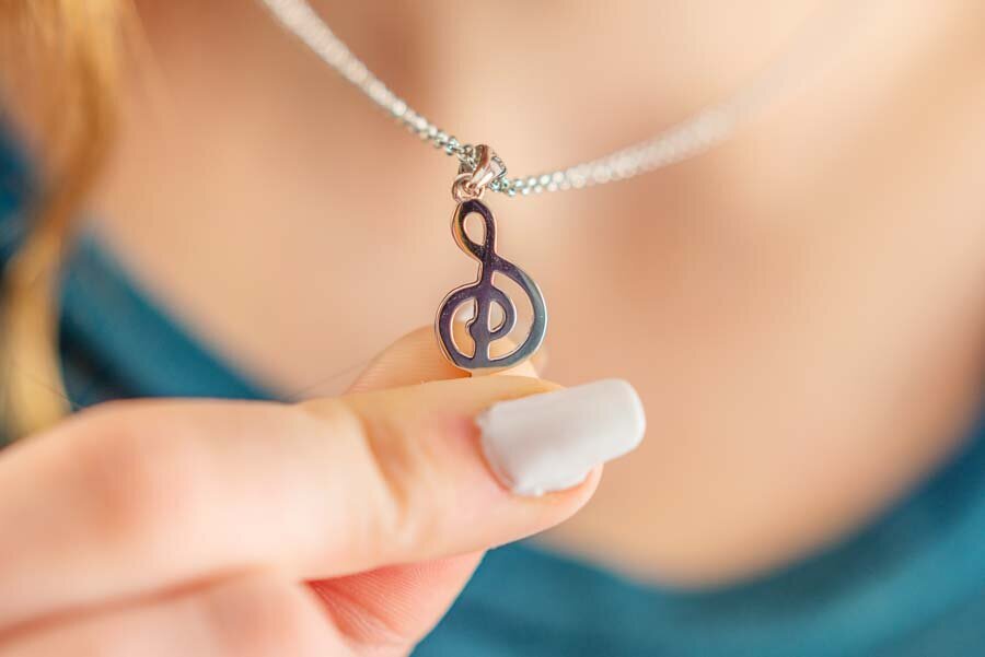 Woman holding a treble clef necklace pendant near her neck, with a focus on the silver pendant and a blurred background.