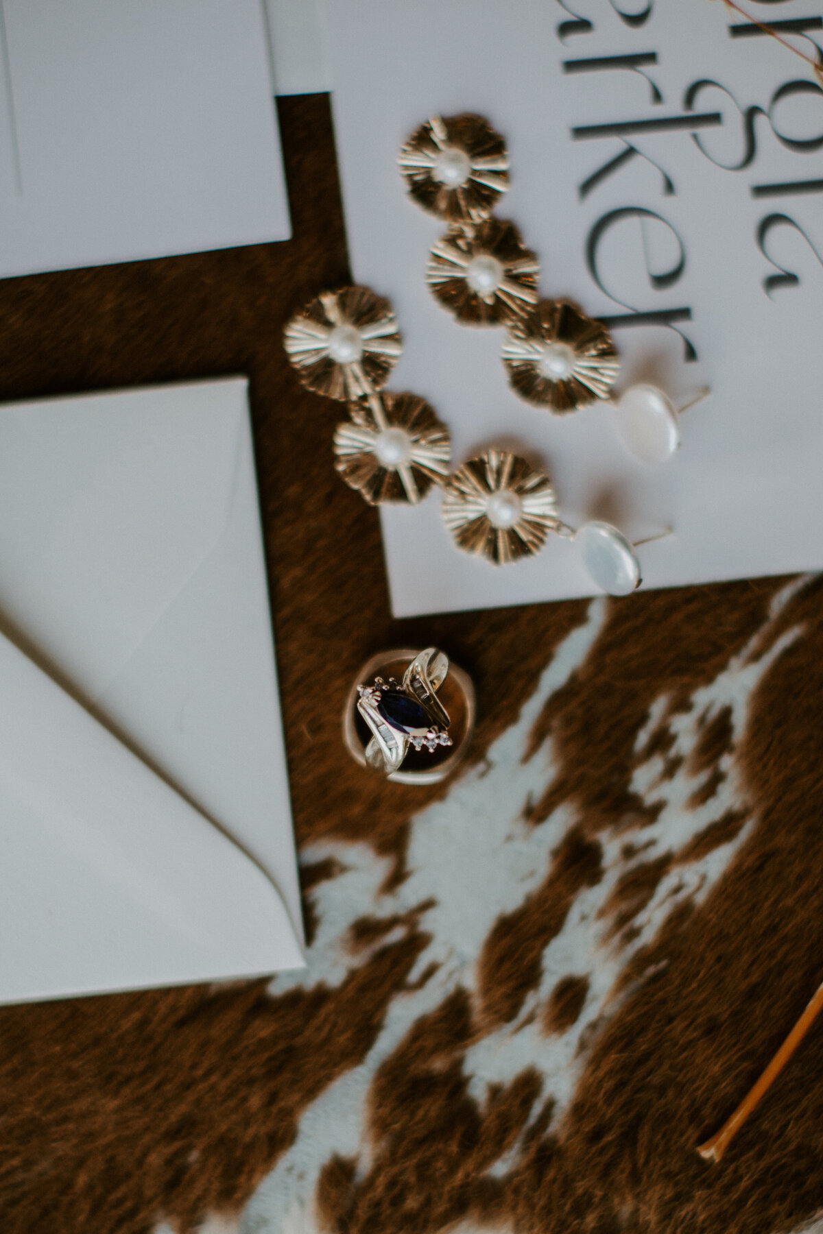 A pair of earrings and wedding bands atop various white wedding stationery with black font on cow hide.