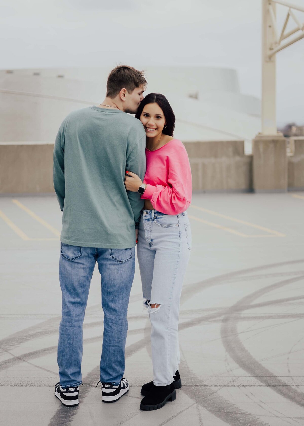 Maddie Rae Photography couple on the top of a parking garage. he is facing away and she is facing the camera holding his arm. he is kissing her head and she is smiling at the camera
