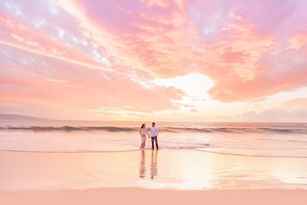 Pregnant couple holds hands at sunset  with beautiful Maui beach behind them