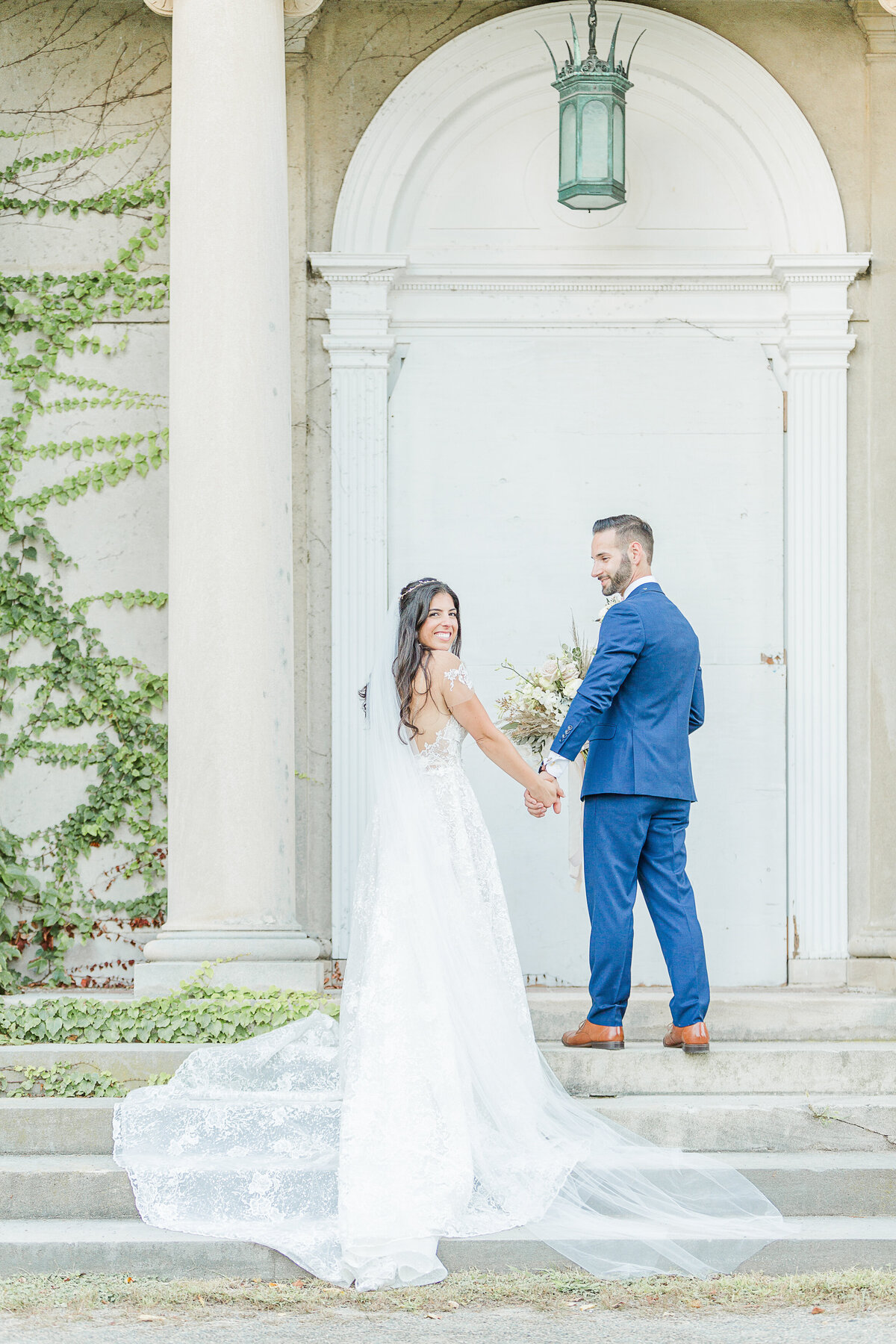 Bride and groom stand on the stone steps at the Eoila Mansion in Connecticut. Their backs are to the camera but are looking over the shoulder so you can see their smiling faces. Captured by New England Wedding Photographer Lia Rose Weddings