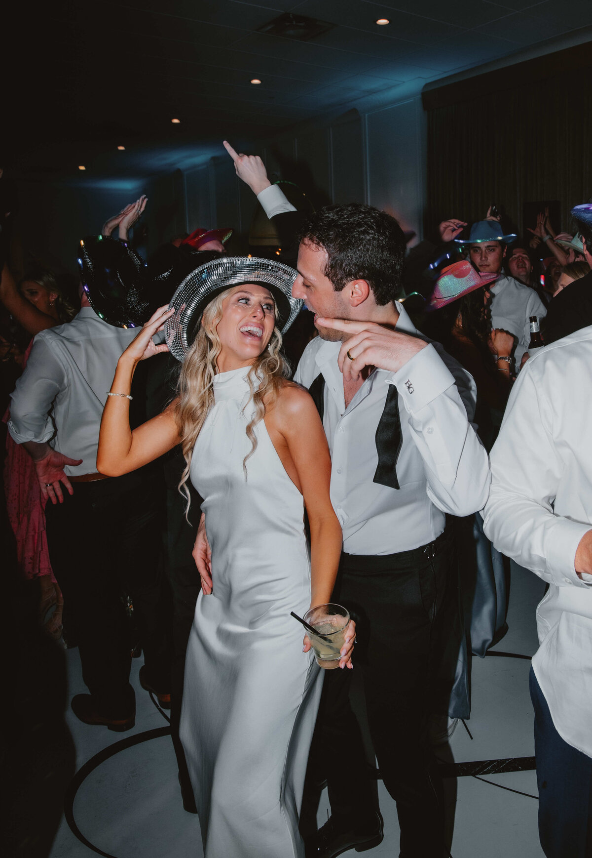 A couple dances joyfully at a party, surrounded by others in formal attire. The woman, wearing a white dress and silver hat, smiles widely.