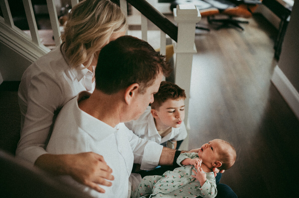 Natural in-home newborn photography in Portland capturing a tender family moment—father cradling newborn while older sibling leans in for a kiss, with mother gently embracing them on the staircase.
