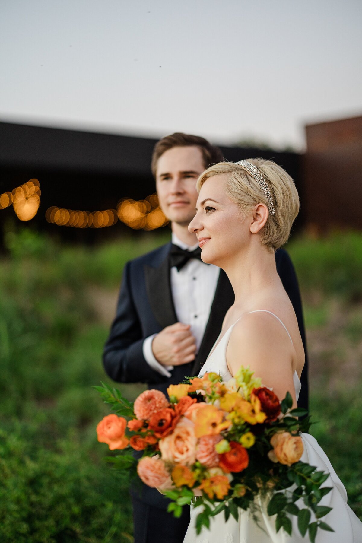 Portrait of a bride and groom looking off into the distance on their wedding day in Dallas, Texas. The bride stands in the foreground and is wearing a sleeveless, long, white dress and is holding a bouquet of orange flowers. The groom stands behind her and is wearing a black tuxedo with a bowtie.