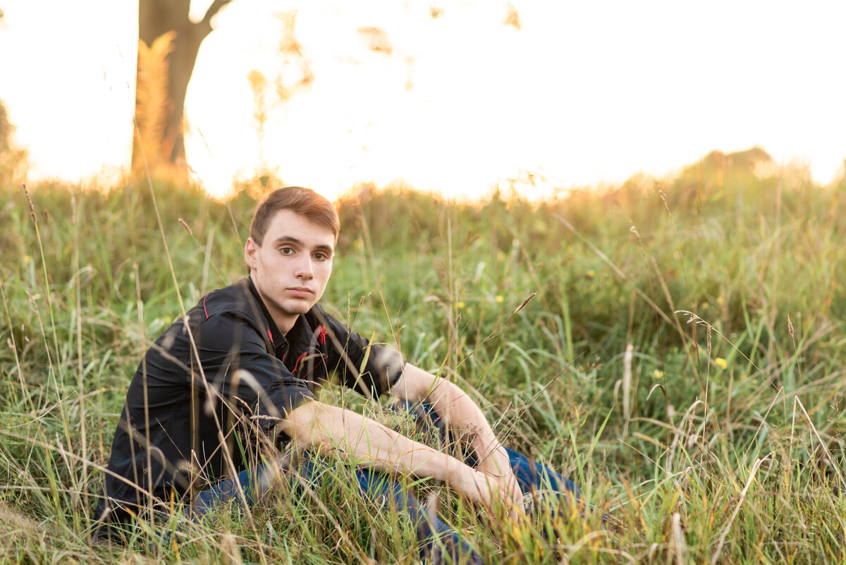 Senior boy sitting in field at sunset, looking at the camera with no smile
