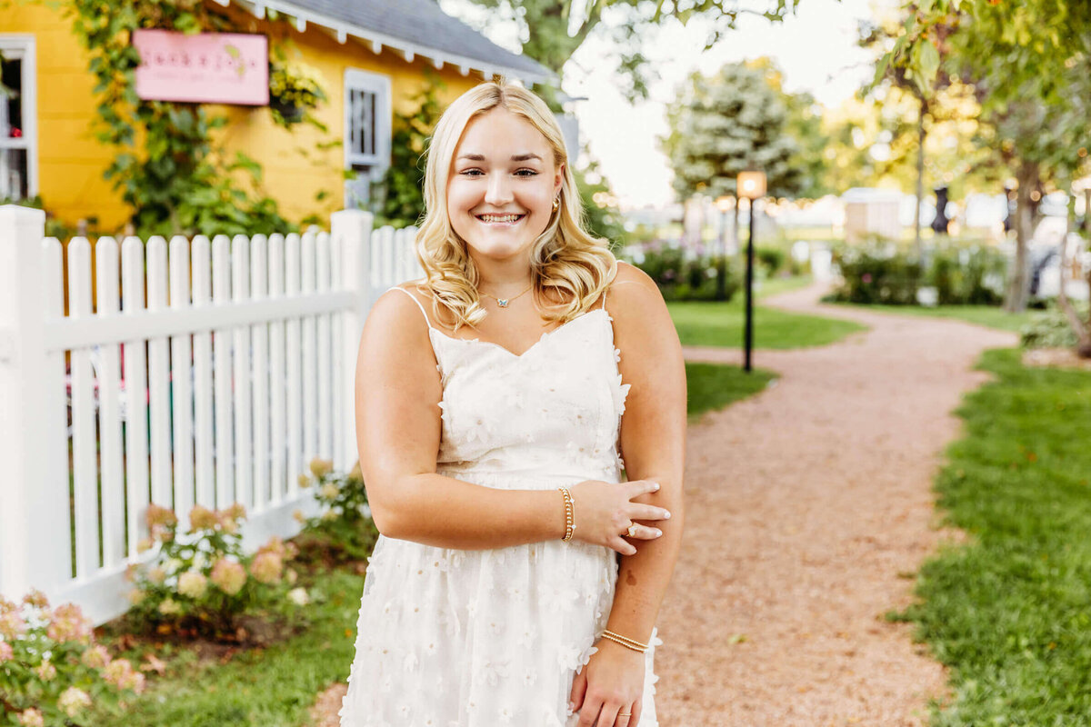 beautiful teenage girl in a white flower dress standing on a pathway by colorful cottages by Ashley Kalbus Photography