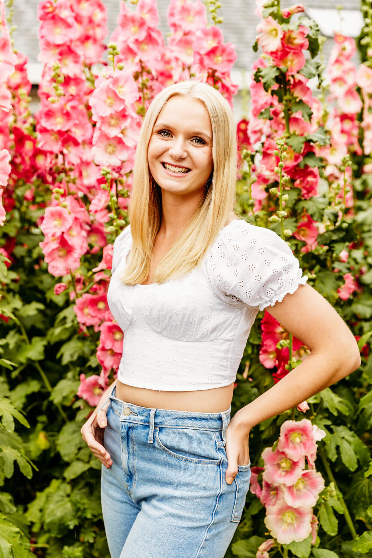 high school senior girl in a white crop top and jeans  standing in a garden of pink holly hocks in Door County captured by Ashley Kalbus Photography
