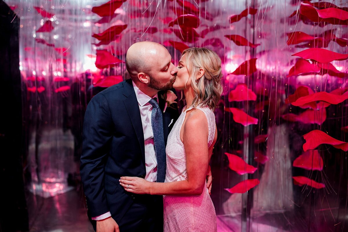 A bride wearing a sleeveless sheath dress kissing a groom in a blue suit at The Hidden Bar at Noelle Hotel Nashville with a mirrored background and hanging red flowers