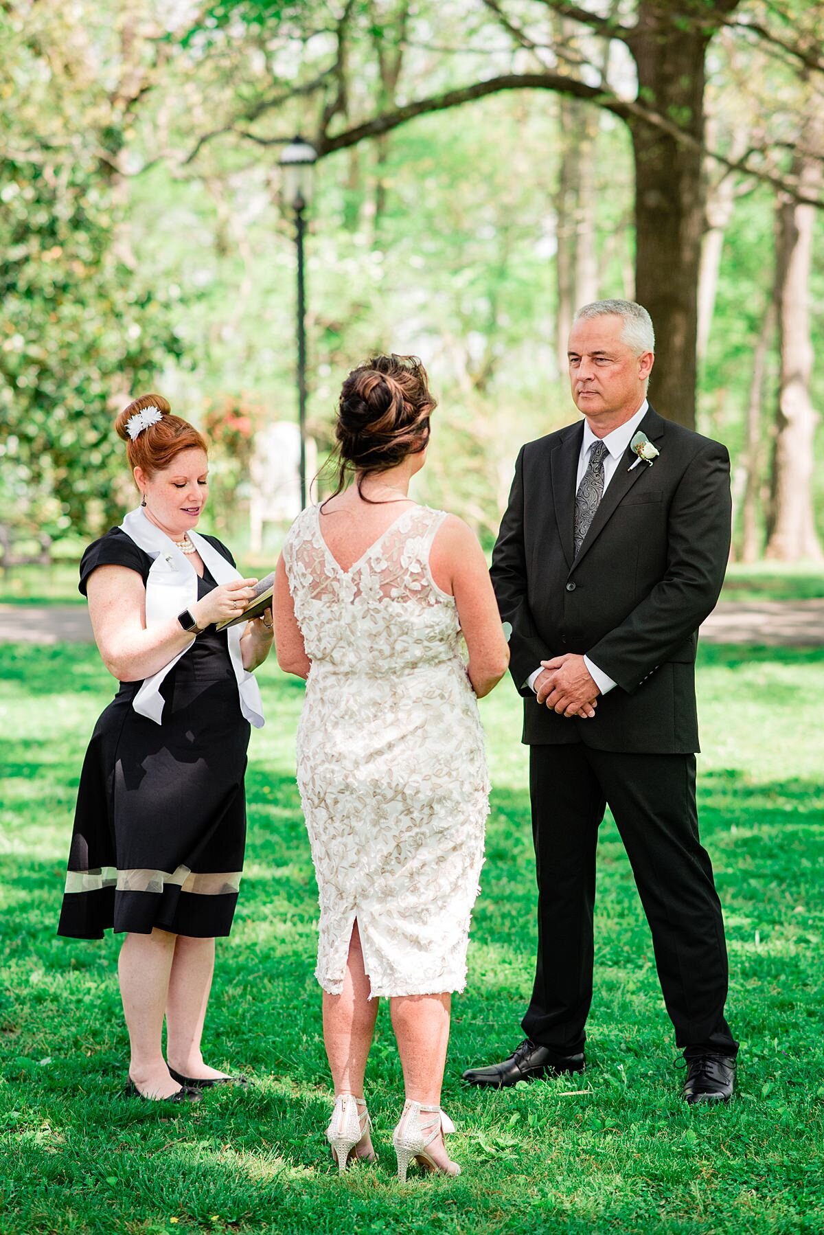 The officiant stands before the bride and groom conducting the wedding ceremony. She is wearing a black dress and a white officiant sash that blows in the breeze.  She has a small gray flower in her hair which is up in a structured bun. The bride is facing away from the camera. She is wearing a sleeveless knee length white lace sheath dress and stilettos with ankle bows. The groom is looking at the bride with his hands clasped in front of him. He is wearing a black suit with a white shirt and gray tie. HE has a white boutonniere on his lapel. They are standing in a green field with a lush wooded area behind them.