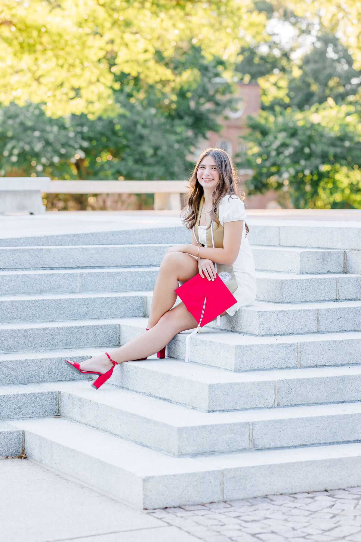 A person in a white dress and red shoes sits on stone steps, holding a red graduation cap, perhaps captured by a skilled senior portrait photographer. In the background, green trees frame a large stone structure, offering a picturesque Fayetteville, NC setting.