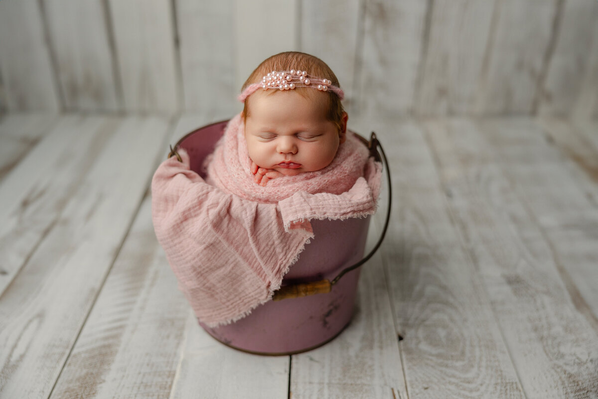 Newborn baby girl swaddled in pink and posed inside a rustic mauve bucket, wearing a delicate pearl headband on a light wood floor backdrop.
Photograph by Inland Empire newborn photographer Kaitlyn Dawn Photography.