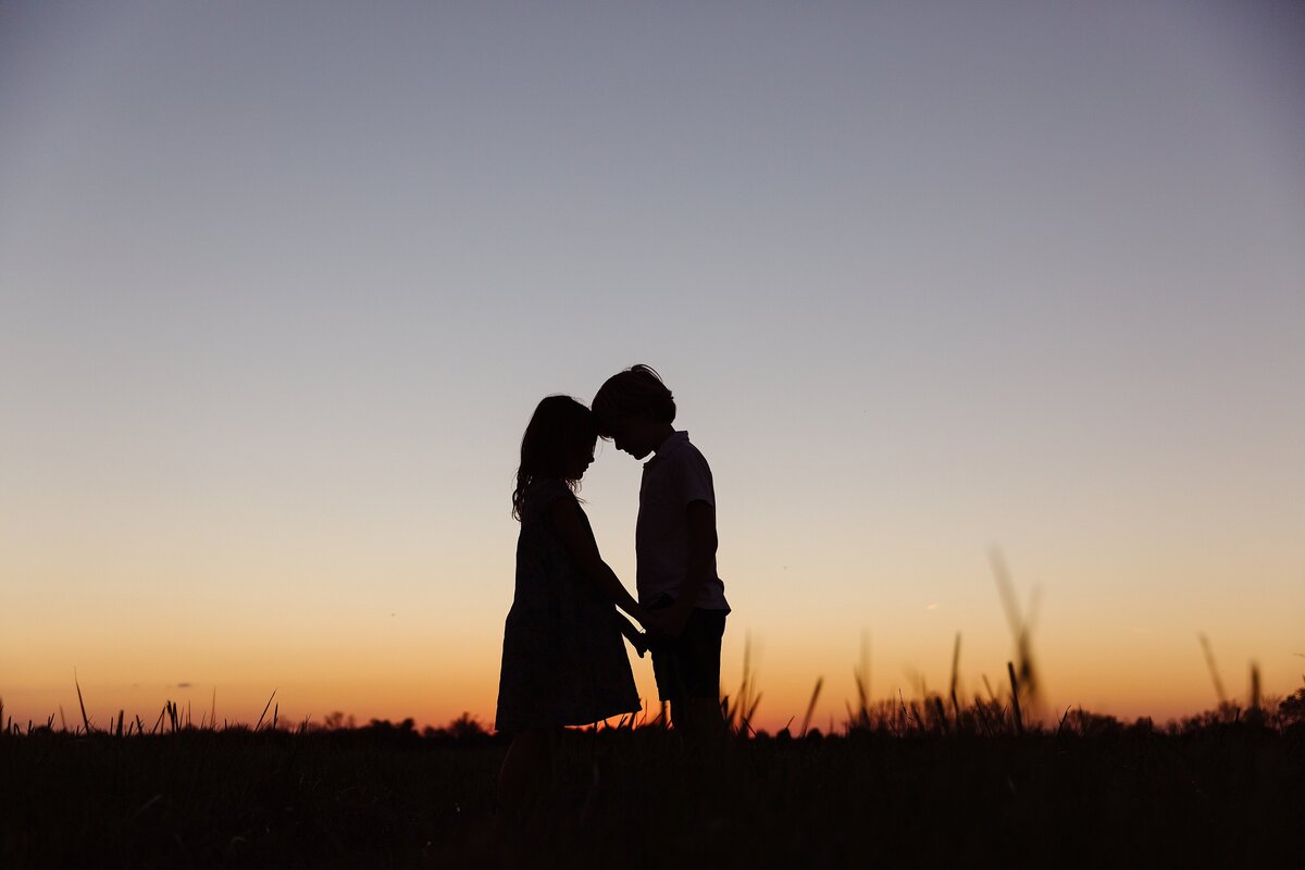 A sunset silhouette image of a brother and sister with heads together.