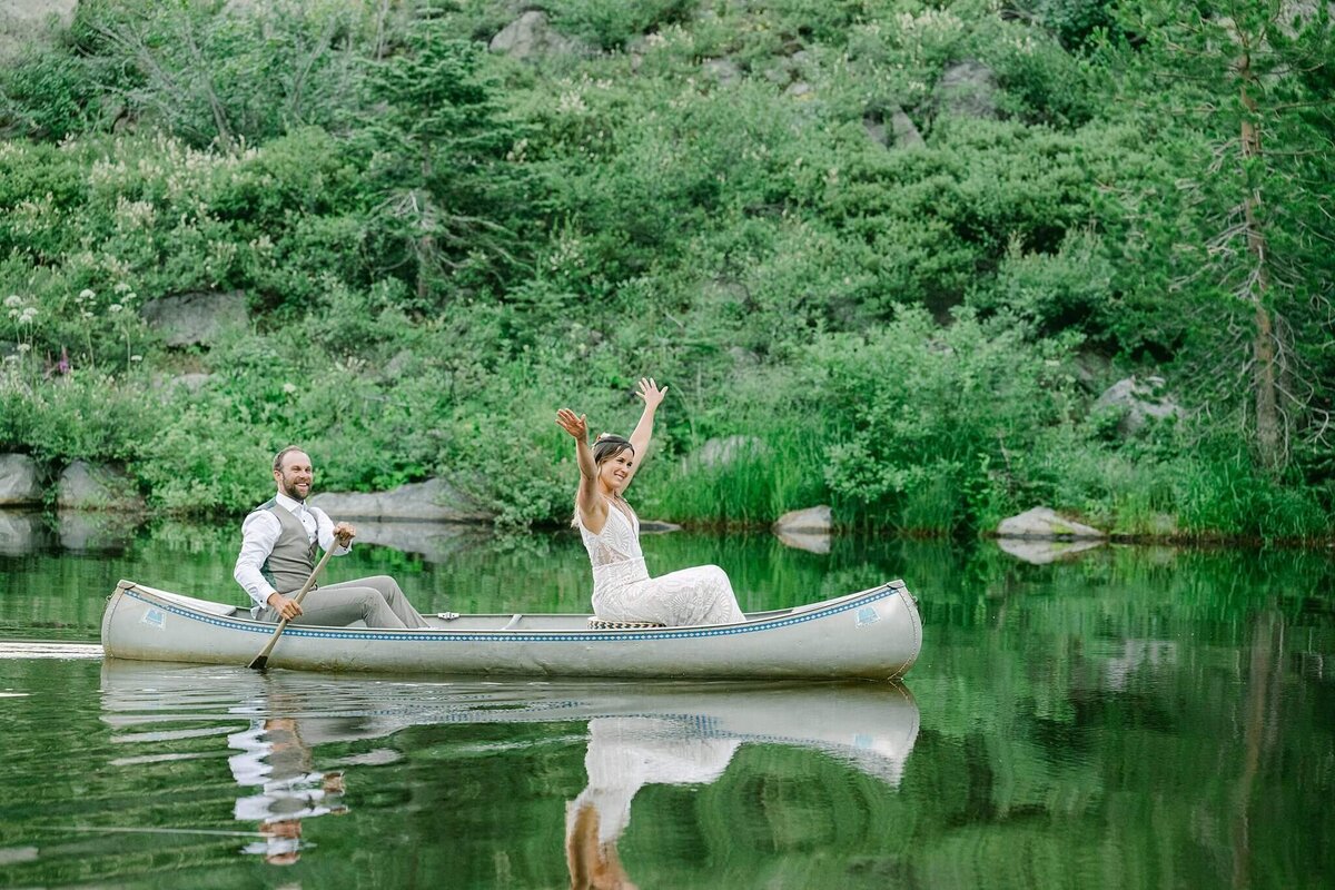 Newlyweds in a canoe waving at their wedding party groom with paddle in pond White Wolf