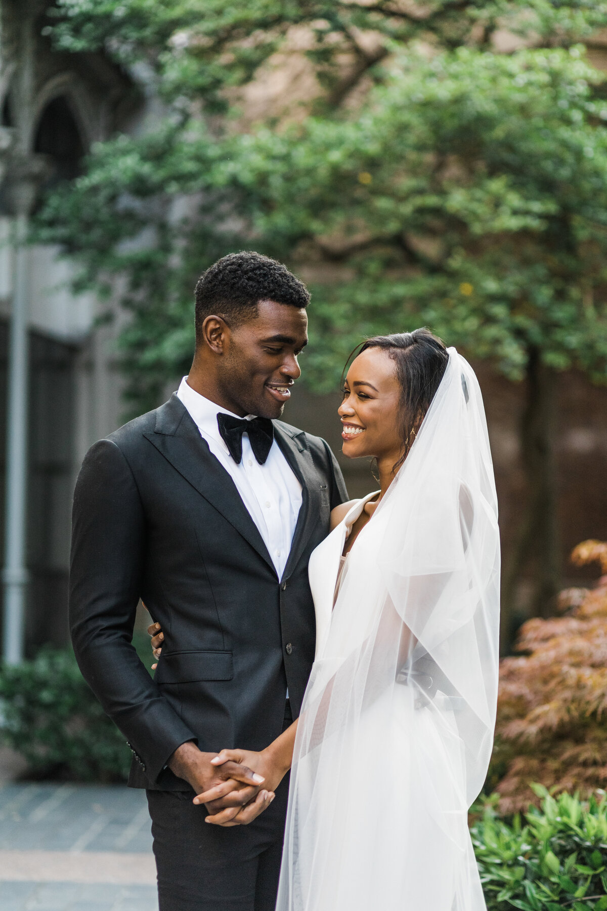 A portrait of a bride and groom posing joyfully together on their wedding day at the Hotel Crescent Court in Dallas, Texas. The bride is on the right and is wearing an elegant white dress with a long flowing veil. The groom is on the left and is wearing a tuxedo with a black bowtie. They are both holding each other close and holding hands.