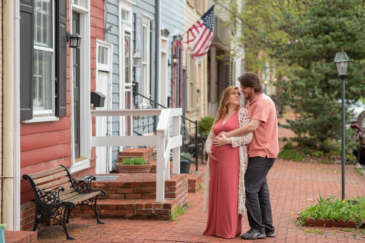 Husband & Pregnant wife smiling at each other on brick street with colorful homes