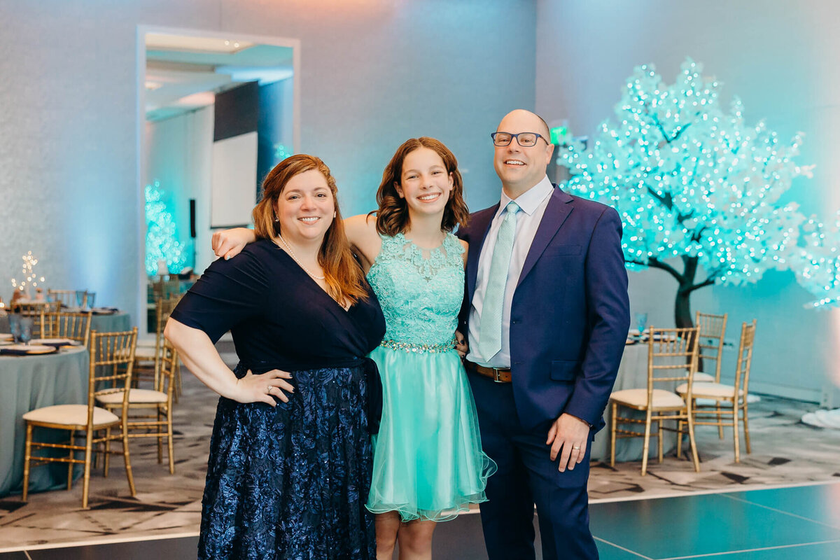 A mother and father dressed in blue suit and dress stand in a ballroom with their teenage daughter smiling for some Bellevue Bar and Bat Mitzvah Photography