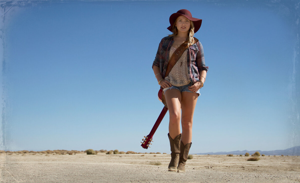 Country music photo Danielle Marie walking through desert wearing red cowboy hat jean shorts with guitar strung across back