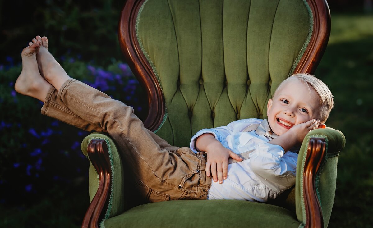 Young boy lays across a victorian green velvet chair and smiles