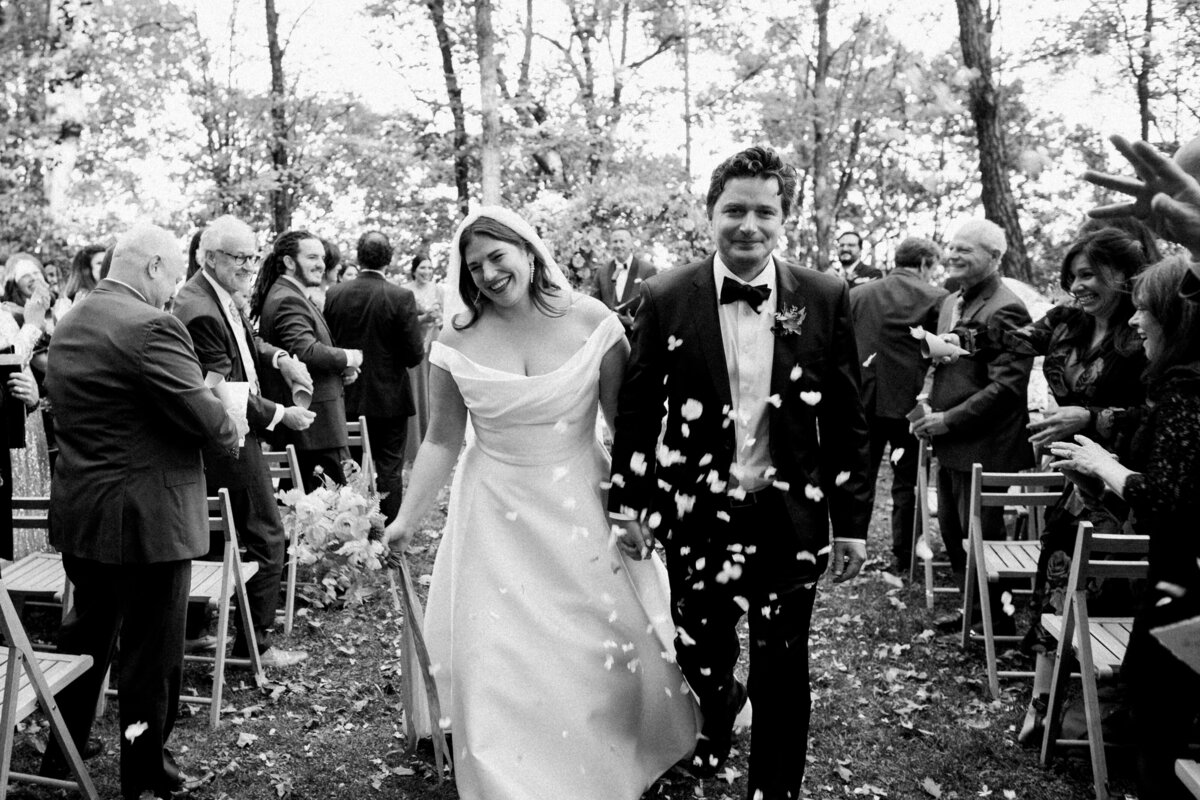 Black and white wedding photo of bride and groom walking down the aisle while guests throw confetti under trees in Hudson Valley