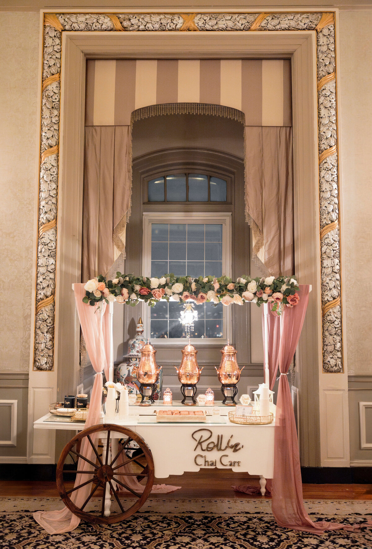 A vintage chai cart decorated with flowers and draped in pink fabric stands in front of a large ornate window. The cart features two copper tea urns and small plates, beneath a sign reading Rollin Chai Cart. The room has elegant decor and patterns.