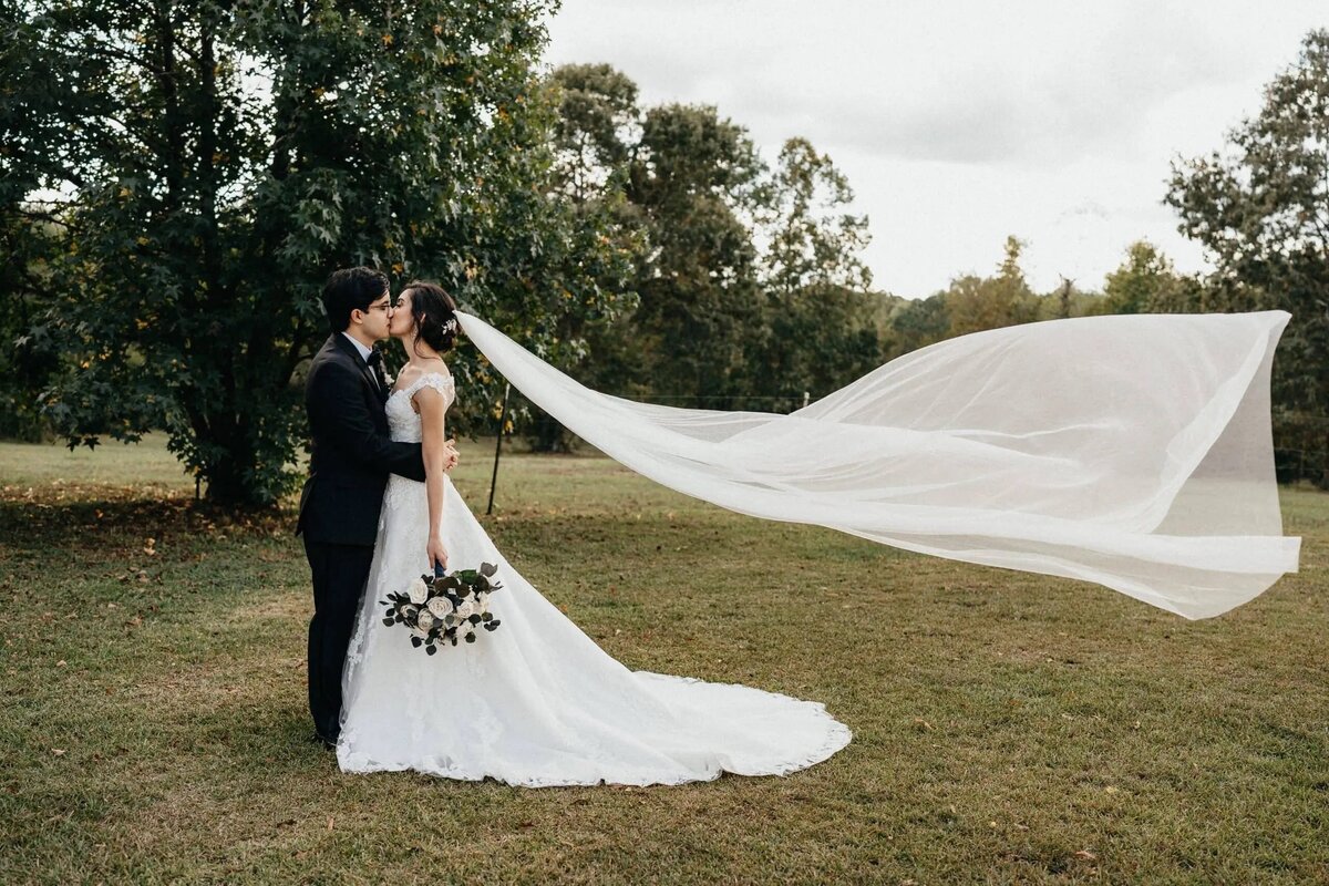 A couple kissing, the bride's veil caught in a breeze, set against a backdrop of greenery, epitomizing a romantic wedding day scene.