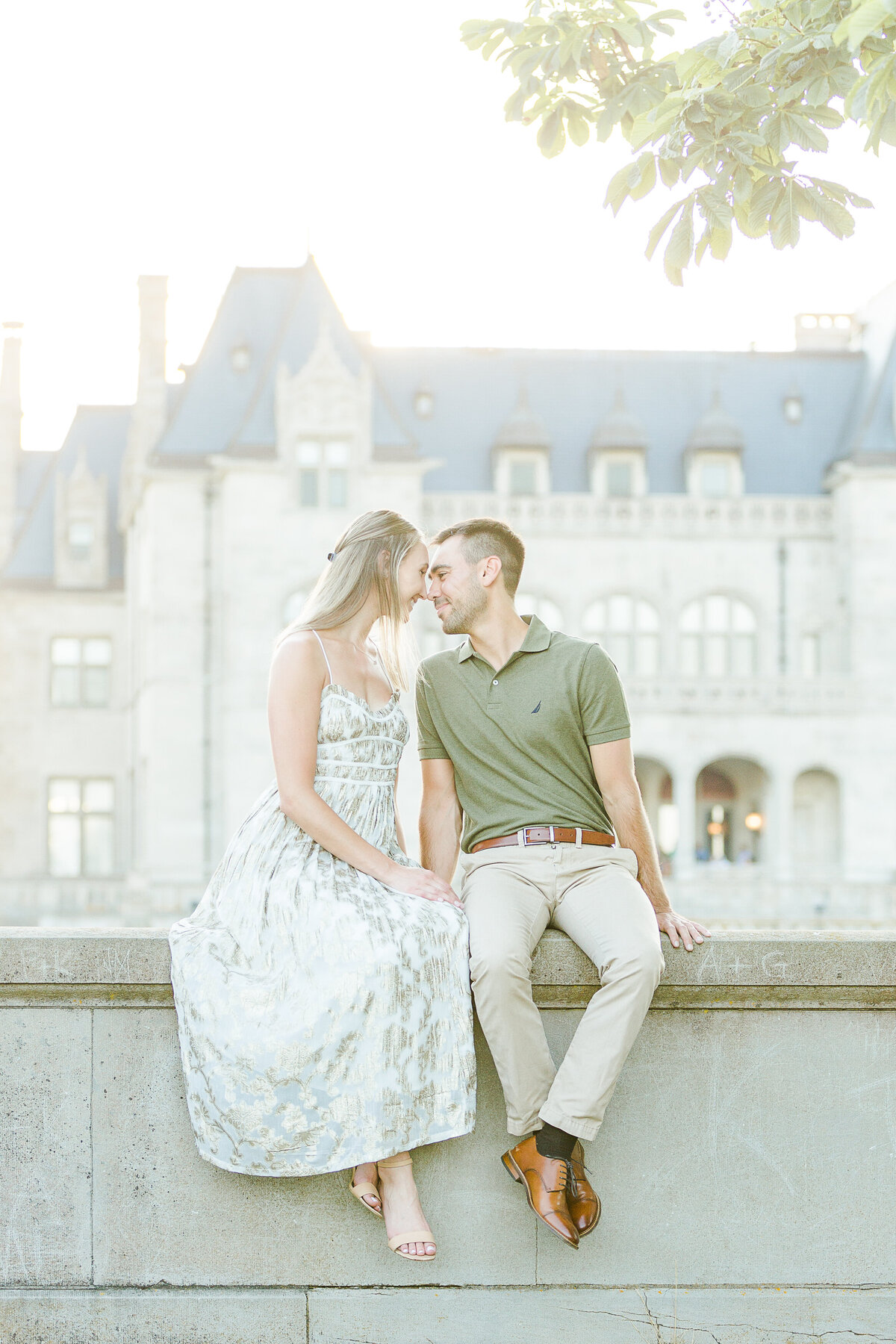 Couple sit beside each other on a stone wall at Salve Regina. Their heads are turned toward each other and their foreheads resting together. Salve Regina's buildings are featured prominently in the background. Captured by Salve Regina wedding photographer Lia Rose Weddings