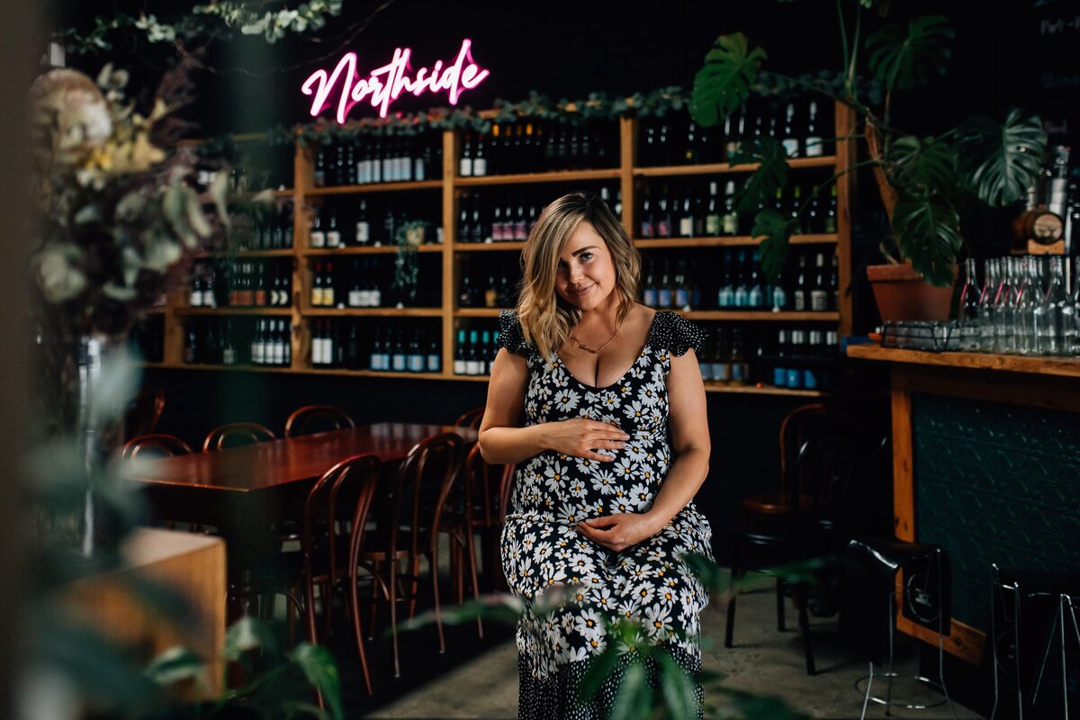 Woman sitting on stool in front of wine rack, holding pregnant belly at Northside Wines, Northcote.