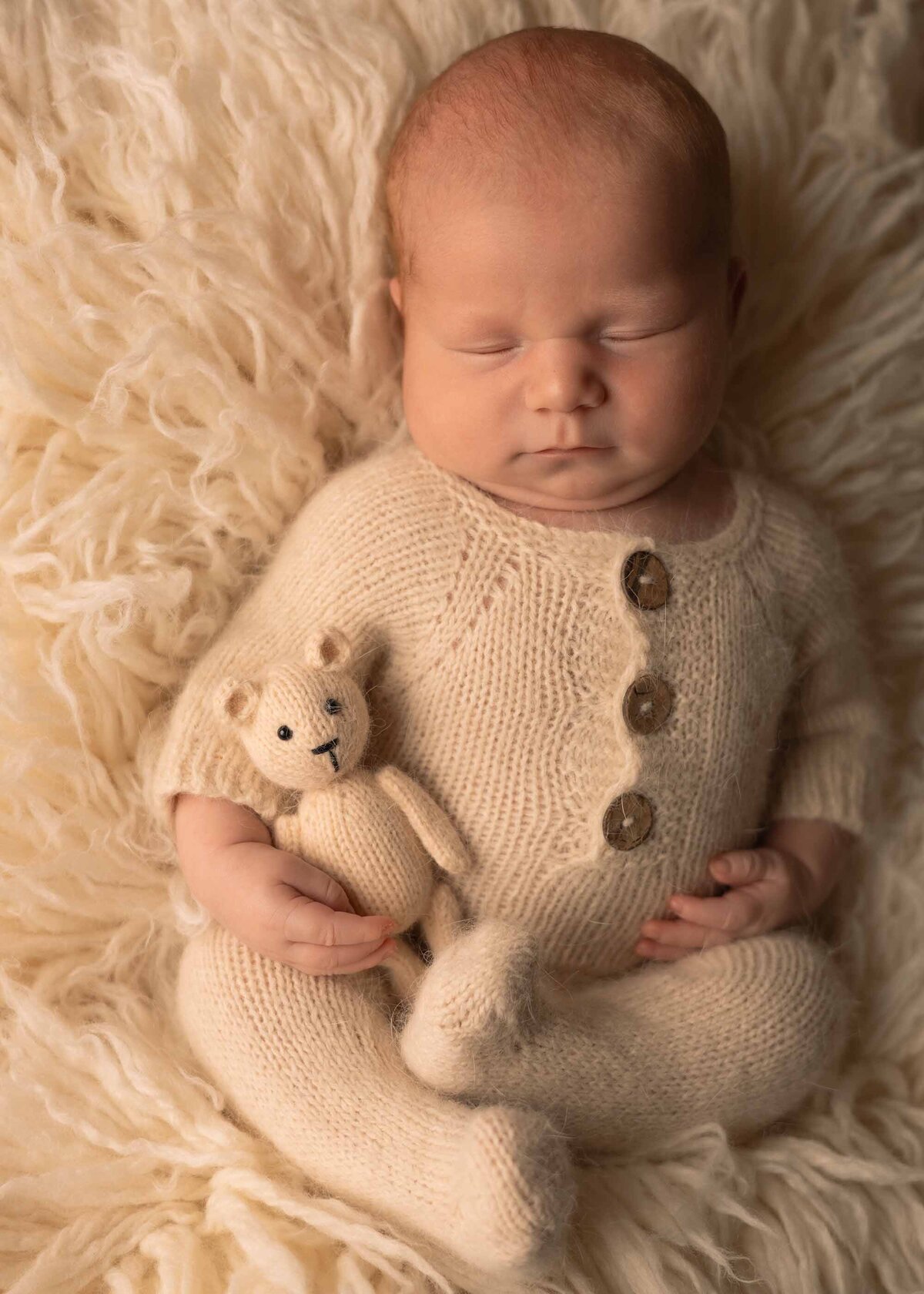 Maddie Rae Photography baby sleeping with his legs crossed. he is wearing a neutral outfit holding a little bear, laying on a neutral rug