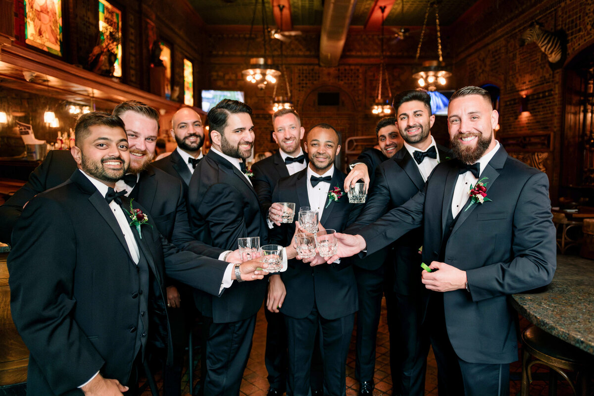 A group of men in formal black suits and bow ties stand together in a warmly lit bar, holding drinks and smiling. The setting features wooden decor and chandeliers, creating a classic, elegant atmosphere.