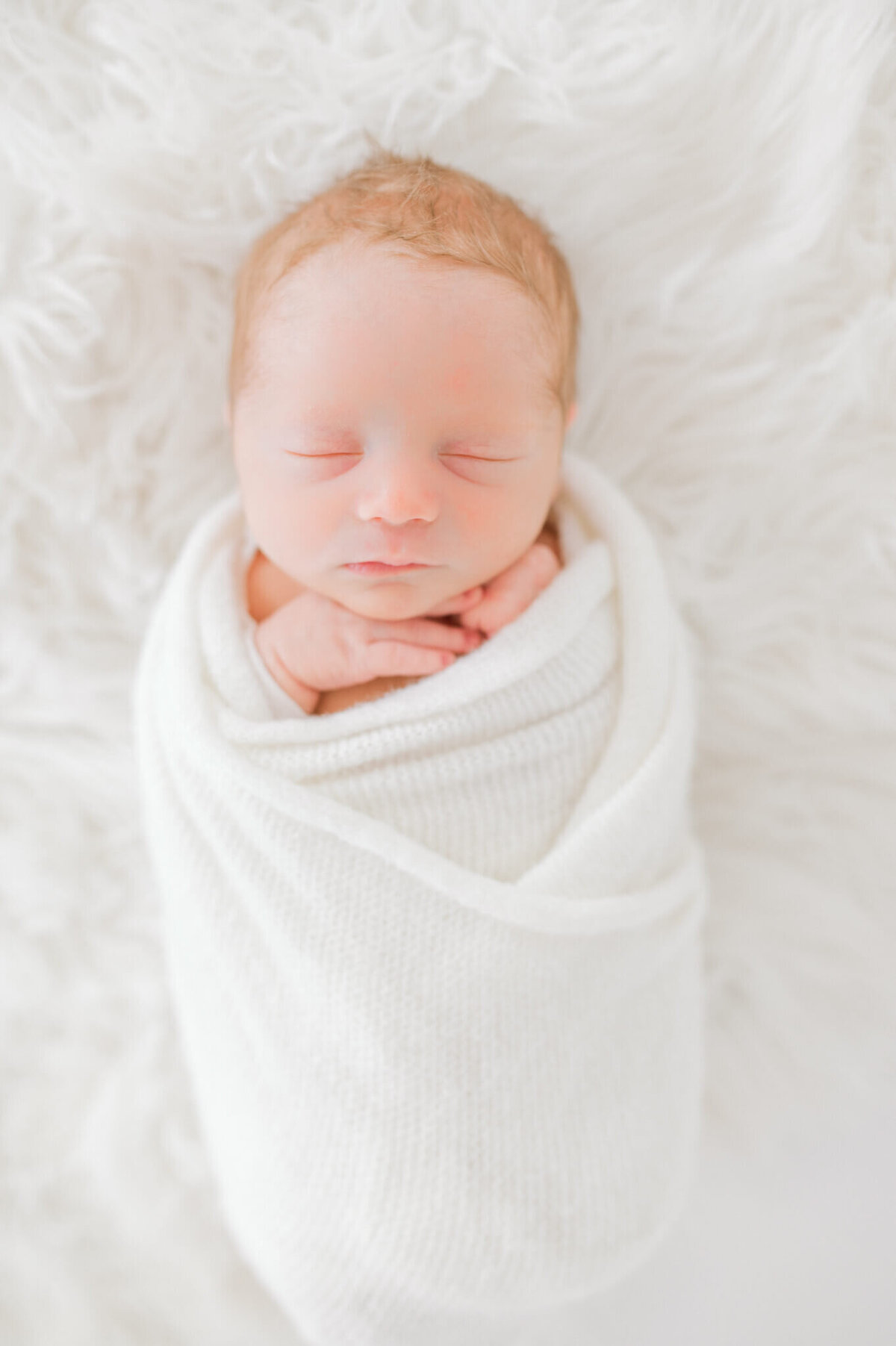 Newborn baby boy with strawberry blonde hair swaddled in wrap on white fur blanket
