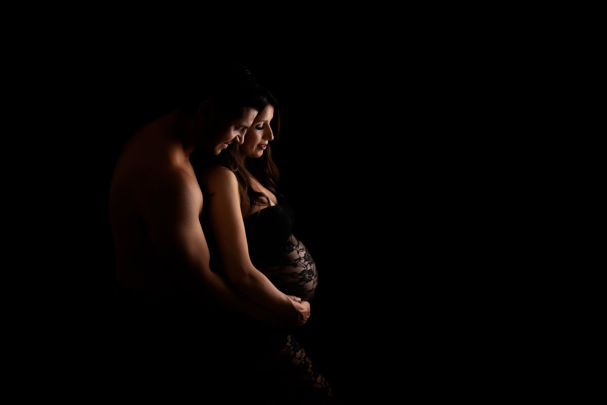 Pregnant woman wearing a fitting black lace dress embraced from behind by her husband on a black background with light catching their faces and her belly.