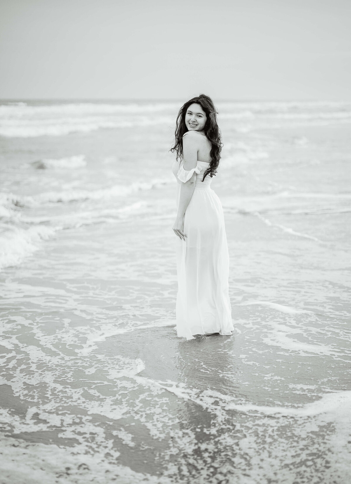 A person with long red hair in a white off-the-shoulder dress stands at the edge of the ocean, with waves gently lapping at their feet on a sandy beach.