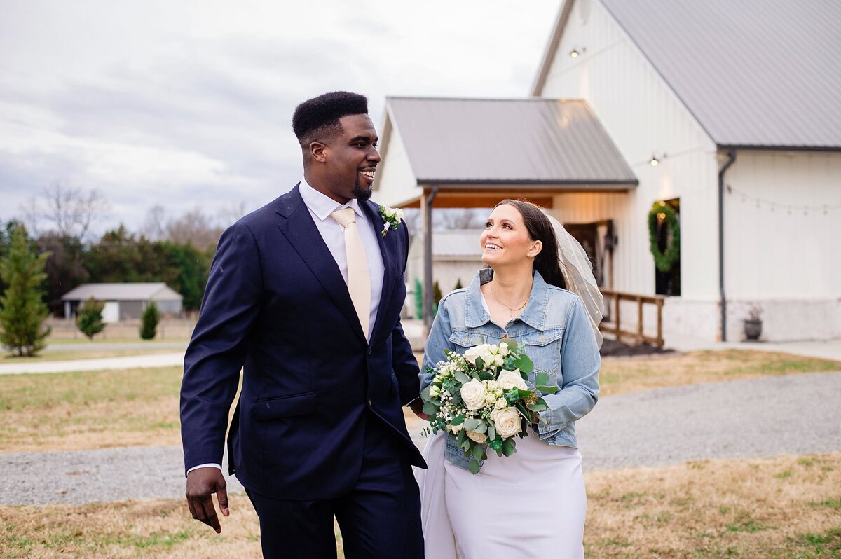A groom wearing a navy suit with a white shirt and ivory tie walks beside the bride who is wearing a short veil and denim jacket with her fitted wedding dress. The bride is holding a large circular bouquet of white roses, white peonies, white hydrangea and greenery at Steel Magnolia Barn.