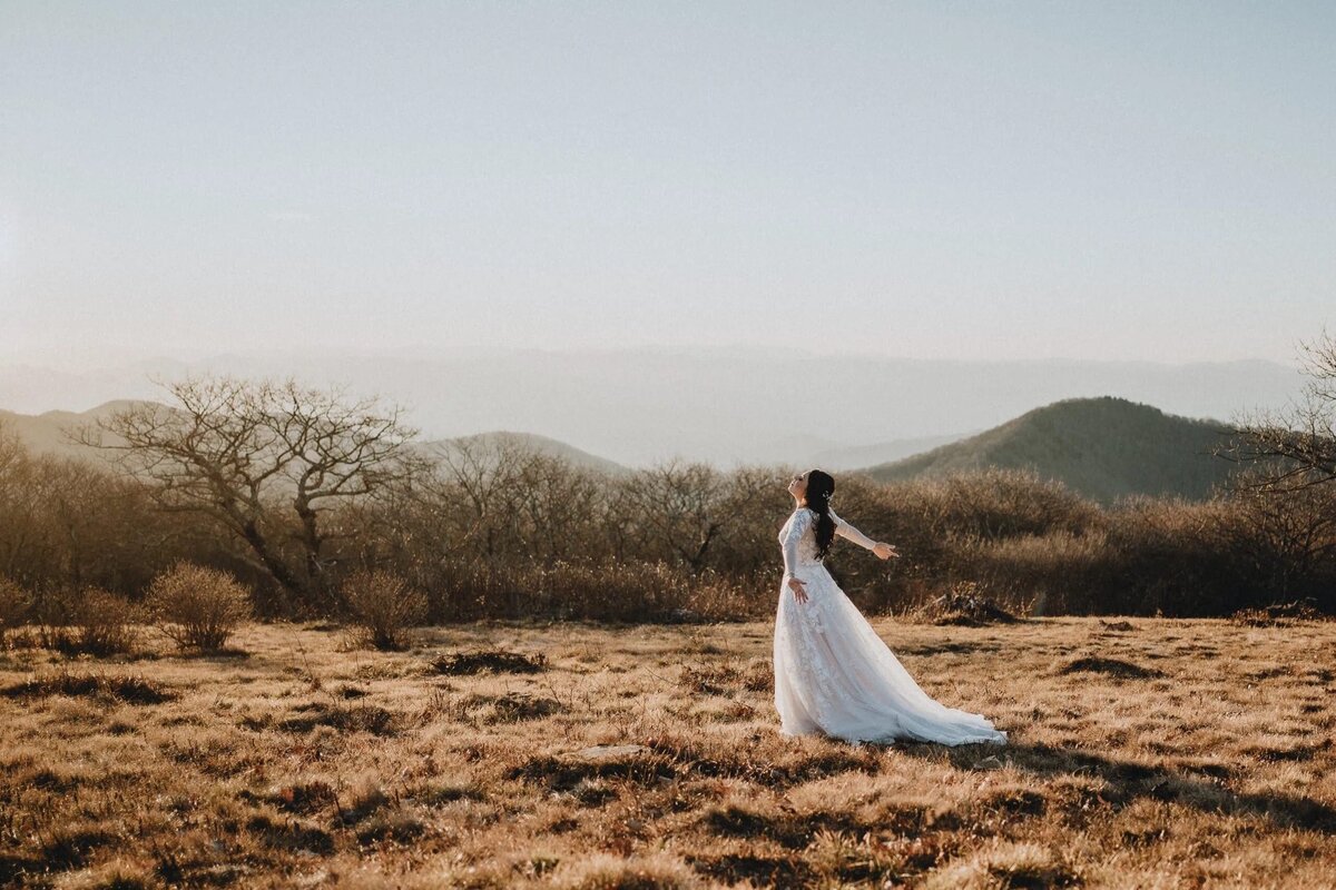 A bride standing alone in a vast field with rolling hills in the background, her wedding dress and veil flowing in the wind