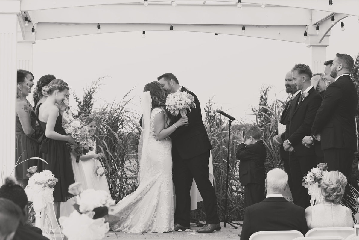 black and white photo of bride and groom first kiss after vow exchange during wedding ceremony at Lombardi's on the Bay