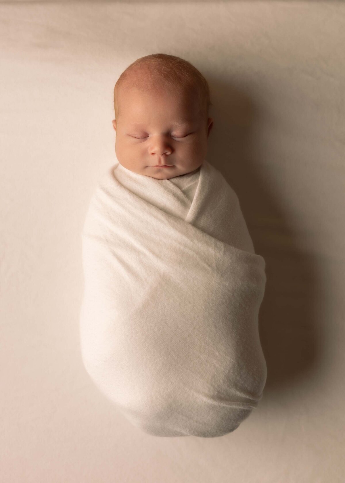 Maddie Rae Photography baby is swaddled up in a white swaddle. he is sleeping and this image is taken from above him