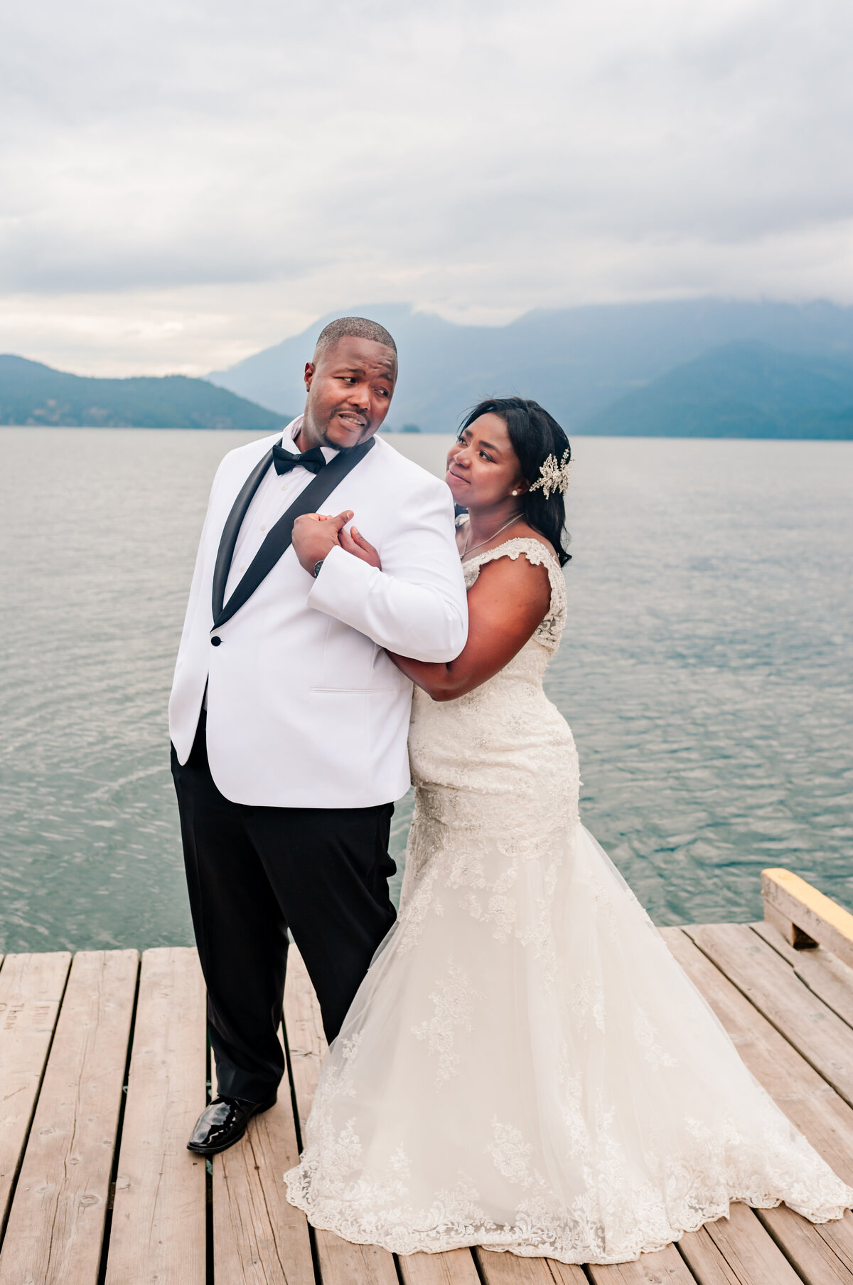 Bride puts chin on the groom shoulder and groom looks over to bride holding her right hand in tight. Back round at Harrison Hot springs lake standing on a dock in white attire. 