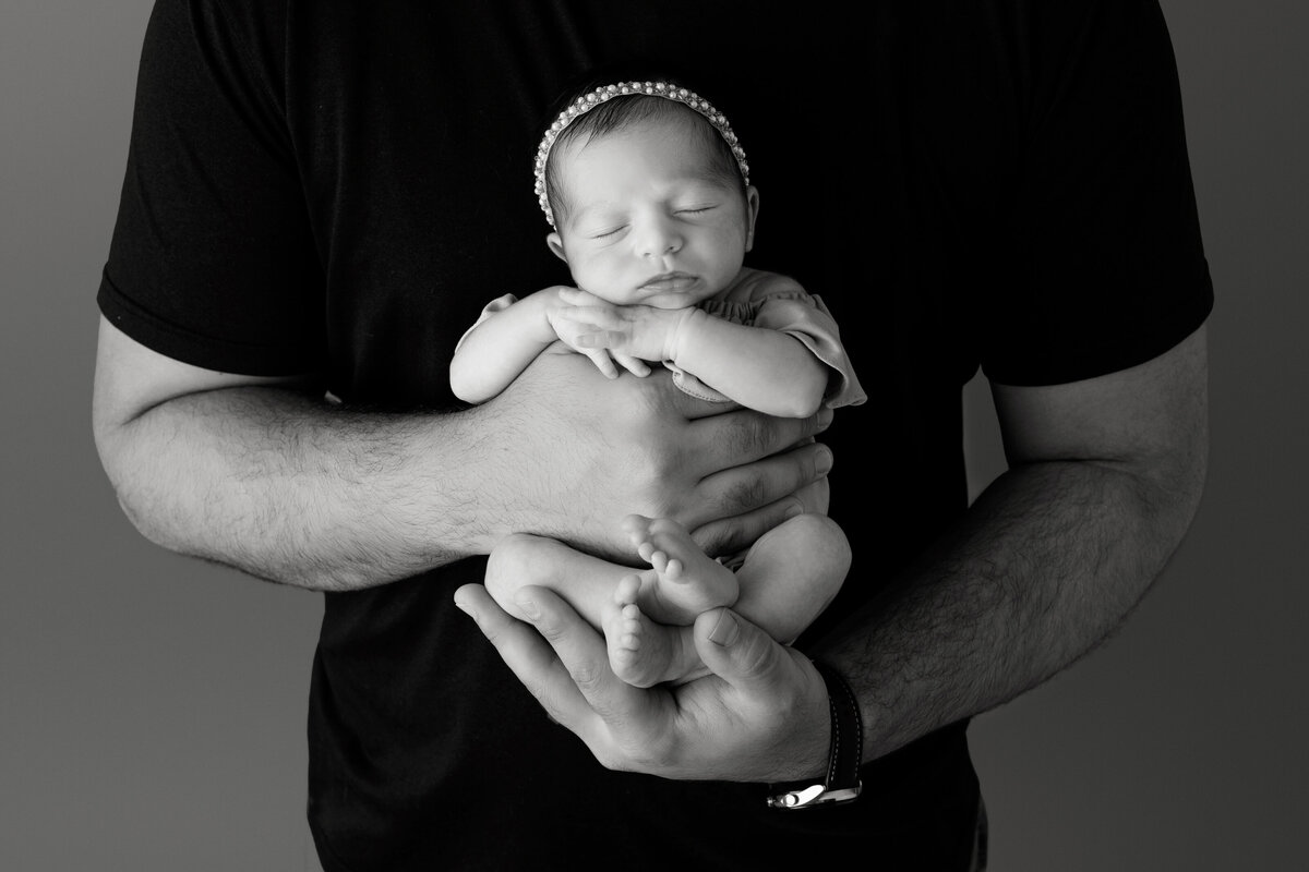 A father in a black shirt cradles his sleeping newborn daughter against his chest in a NJ Newborn Photography studio