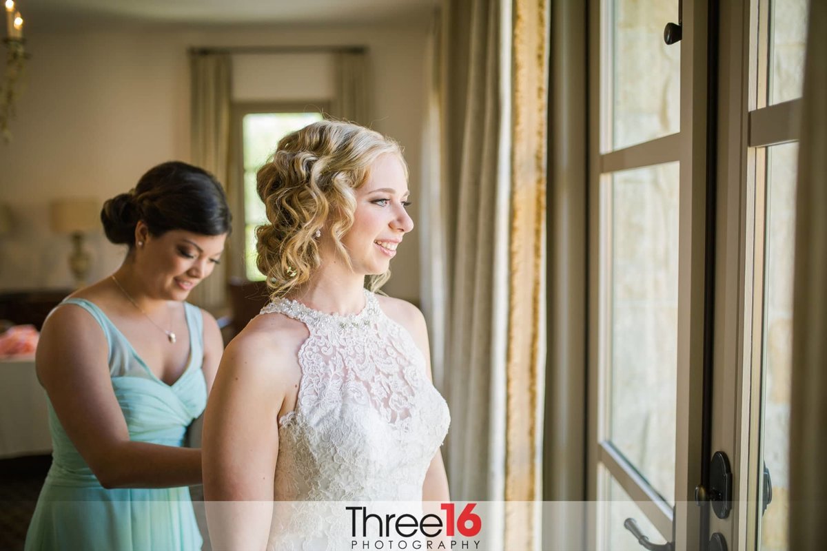 Bride having her dressed buttoned up prior to the ceremony as she looks out the window