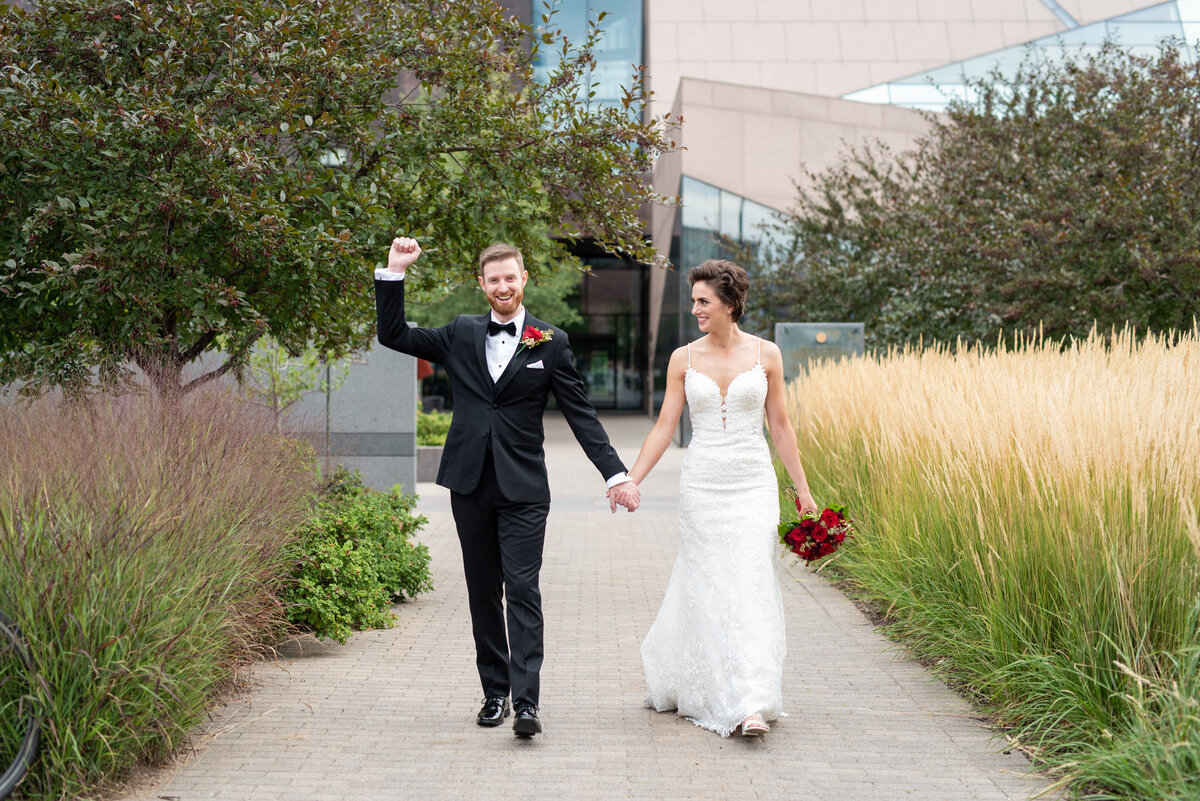 Bride and groom walk down an outdoor path holding hands and smiling while groom cheers and raises his arm in celebration