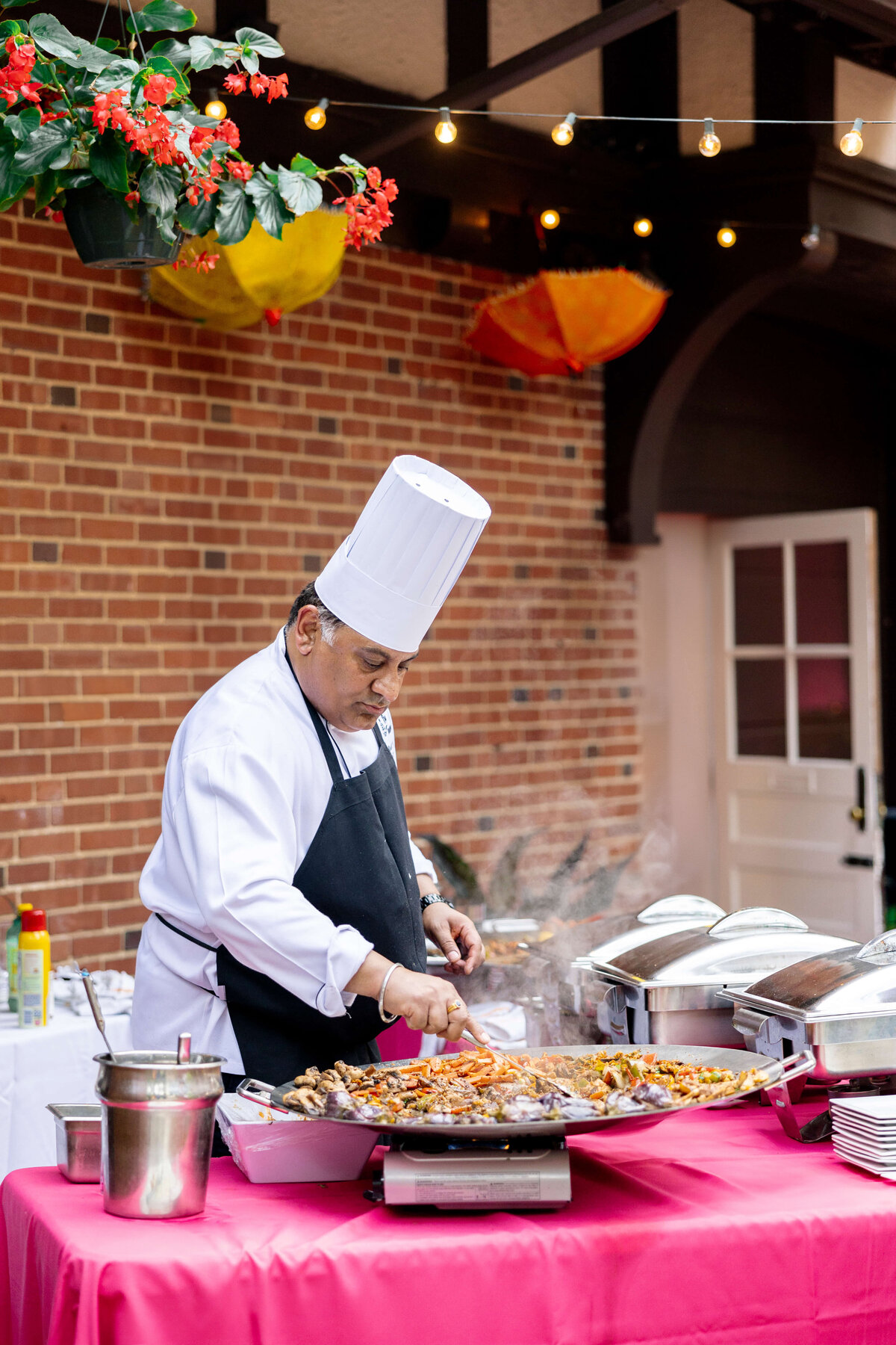 A chef in a white uniform and tall hat is cooking a large pan of paella outdoors. The setup is on a pink tablecloth with various cooking utensils. Overhead string lights and red flowers hang nearby against a brick wall.
