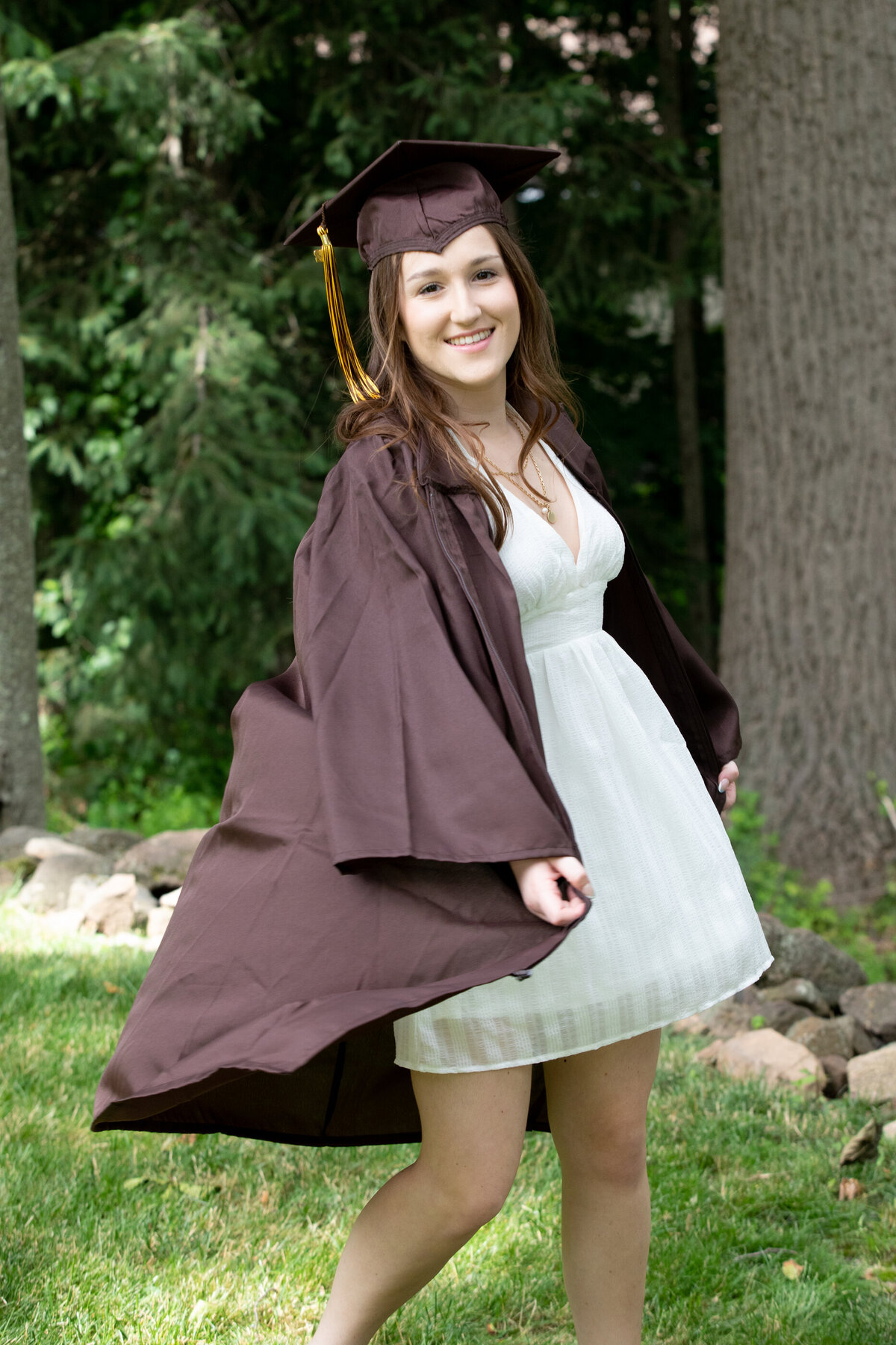 white teenage girl with brown hair twirls in a brown graduation cap and gown and a white dress Clarkstown South senior portrait photographer Milestone Images