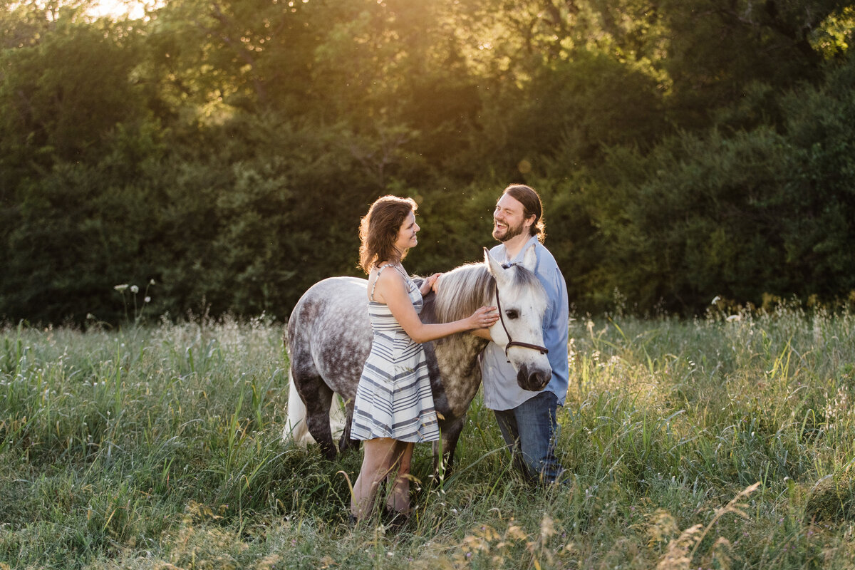 A couple laughing and posing with their horse in a field during their engagement session at White Rock Lake in Dallas, Texas. The woman on the left is wearing a short white dress covered in stripes while the man on the right is wearing a light blue dress shirt and jeans. Their horse is grey and dappled with white spots.