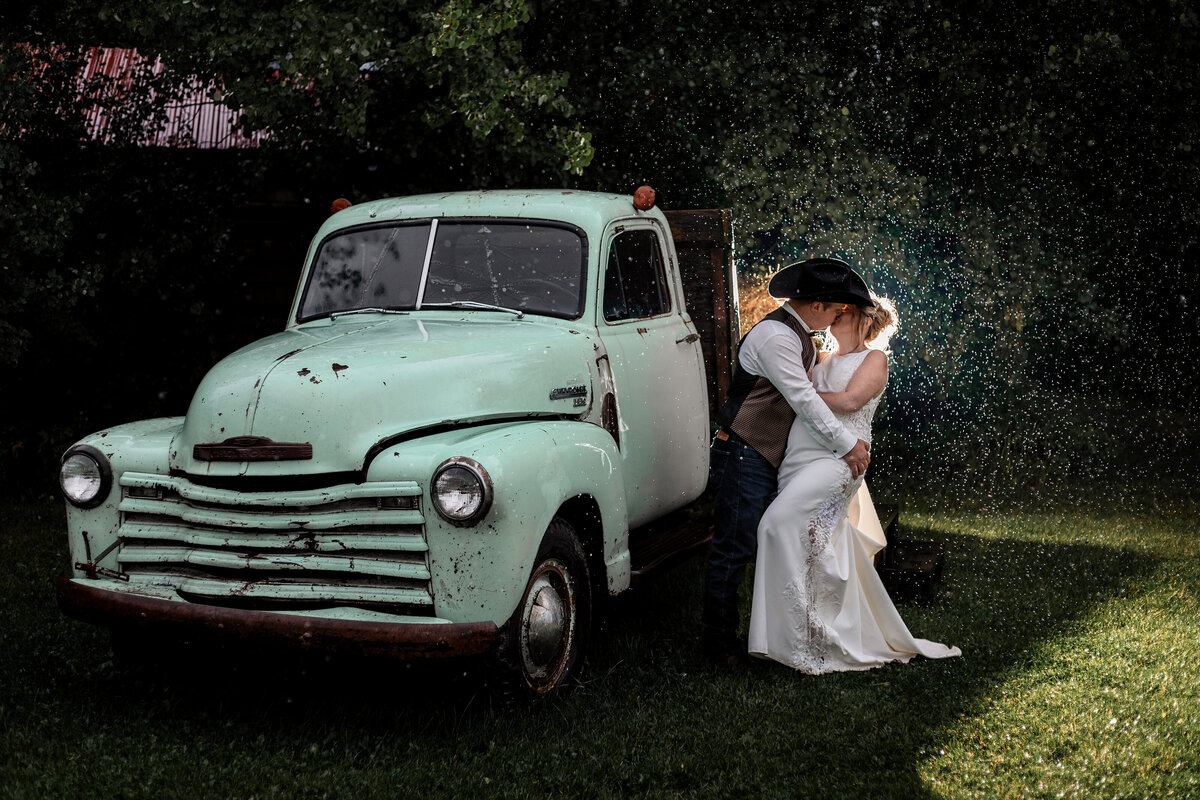 Bride and groom kiss beside an old truck in the rain