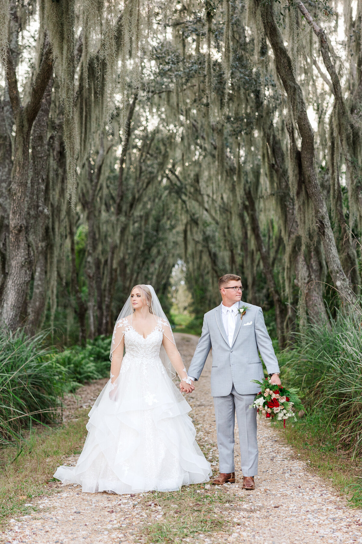 bride and groom hold hands and look in opposite directions as they stand on dirt road