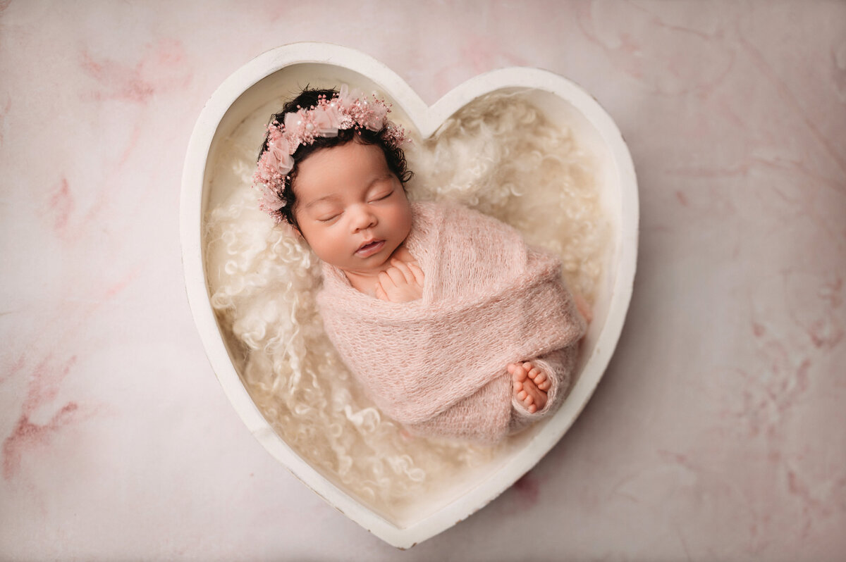 newborn baby girl with curly black hair wrapped in baby pink wrap with her toes sticking out inside a white heart bowl prop sleeping