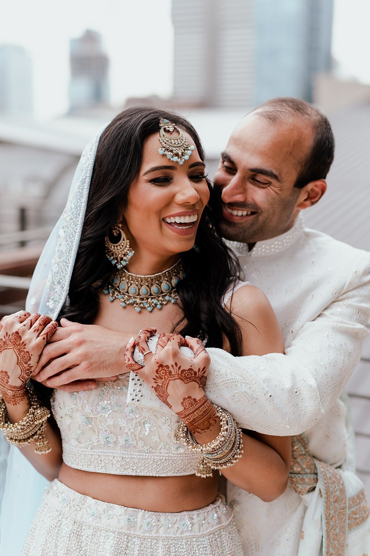 Indian Bride wearing a white beaded saree with a light blue dupatta with gold and light blue Indian wedding jewelry shows off her wedding henna as the groom embraces her. The Indian Groom is wearing a white sherwani with a light blue and gold dupatta. He smiles down at the bride who is laughing.