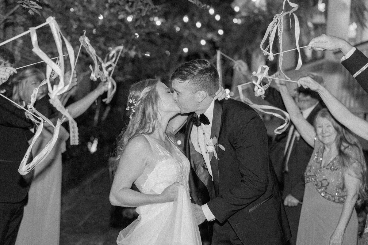 Ribbon stick wedding reception exit. Black and white photo with flash. Bride and groom kiss in the middle of their wedding guests on the way to getaway car at Parsonage Fall wedding. Kailee DiMeglio Photography.